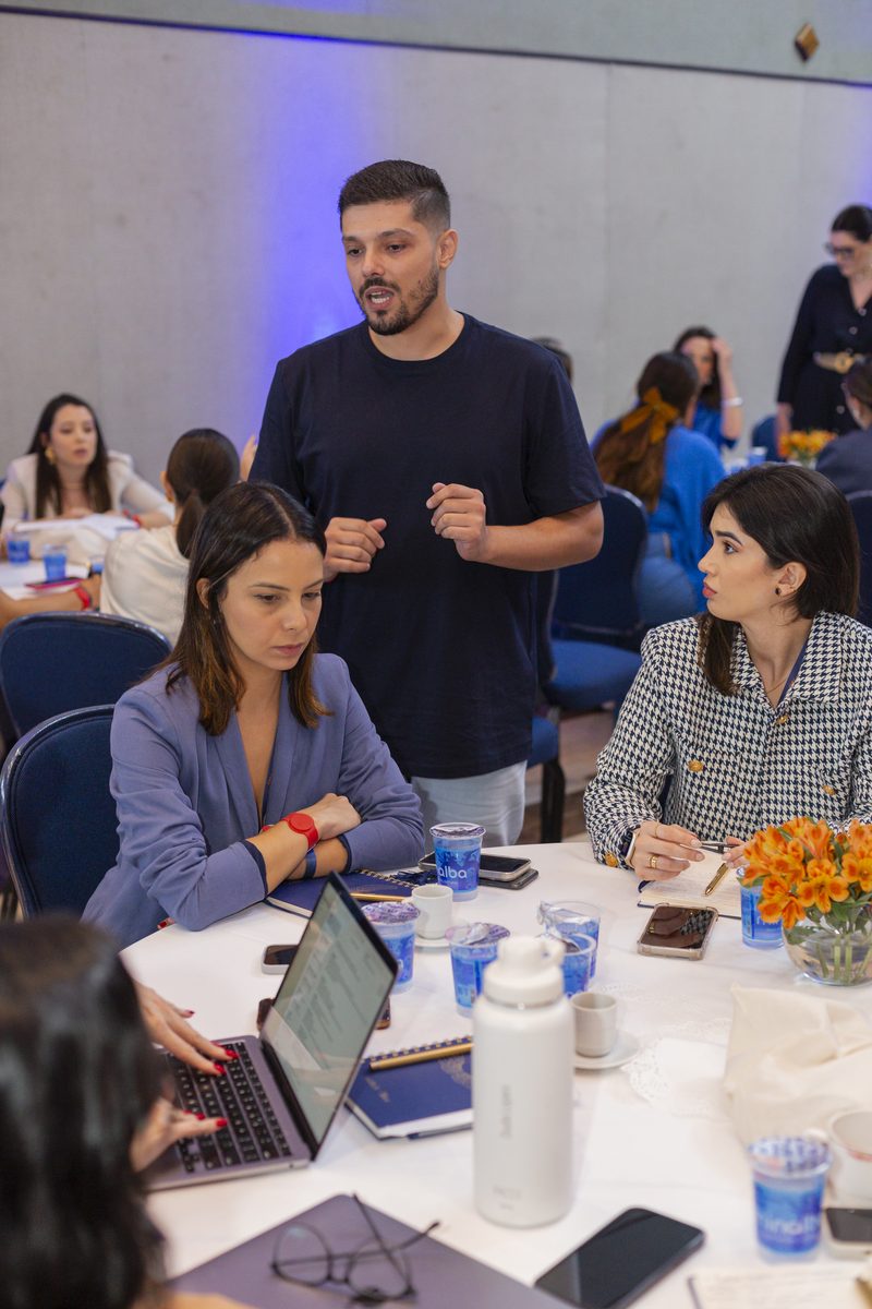 Victor Luporini em pé junto a uma mesa de participantes com computadores abertos, explicando